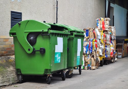 A large, green wheeled rubbish bin with a closed lid positioned on a paved area beside a light-colored exterior wall, with three rectangular white labels on its front. To the right of the bin, there are multiple bundled stacks of flattened cardboard boxes tied with string, and a section of a warehouse or building with large glass windows and a partially visible black door frame in the background. The scene appears to be outdoors on a driveway or street in an urban or residential area, with a concrete surface and minimal surrounding vegetation, reflecting typical waste collection and disposal practices in Nottinghill. The image is relevant to house clearance and rubbish removal services provided by Nottinghill House Clearance, illustrating typical waste materials handled in such operations.